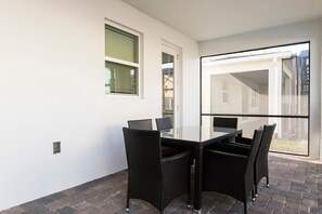 En patio med et glasbord og fletstole, en hvid væg og et vindue med persienner.