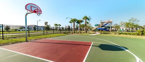 An outdoor basketball court with a red and green playing surface, surrounded by a fence, palm trees, and a water slide structure in the background.