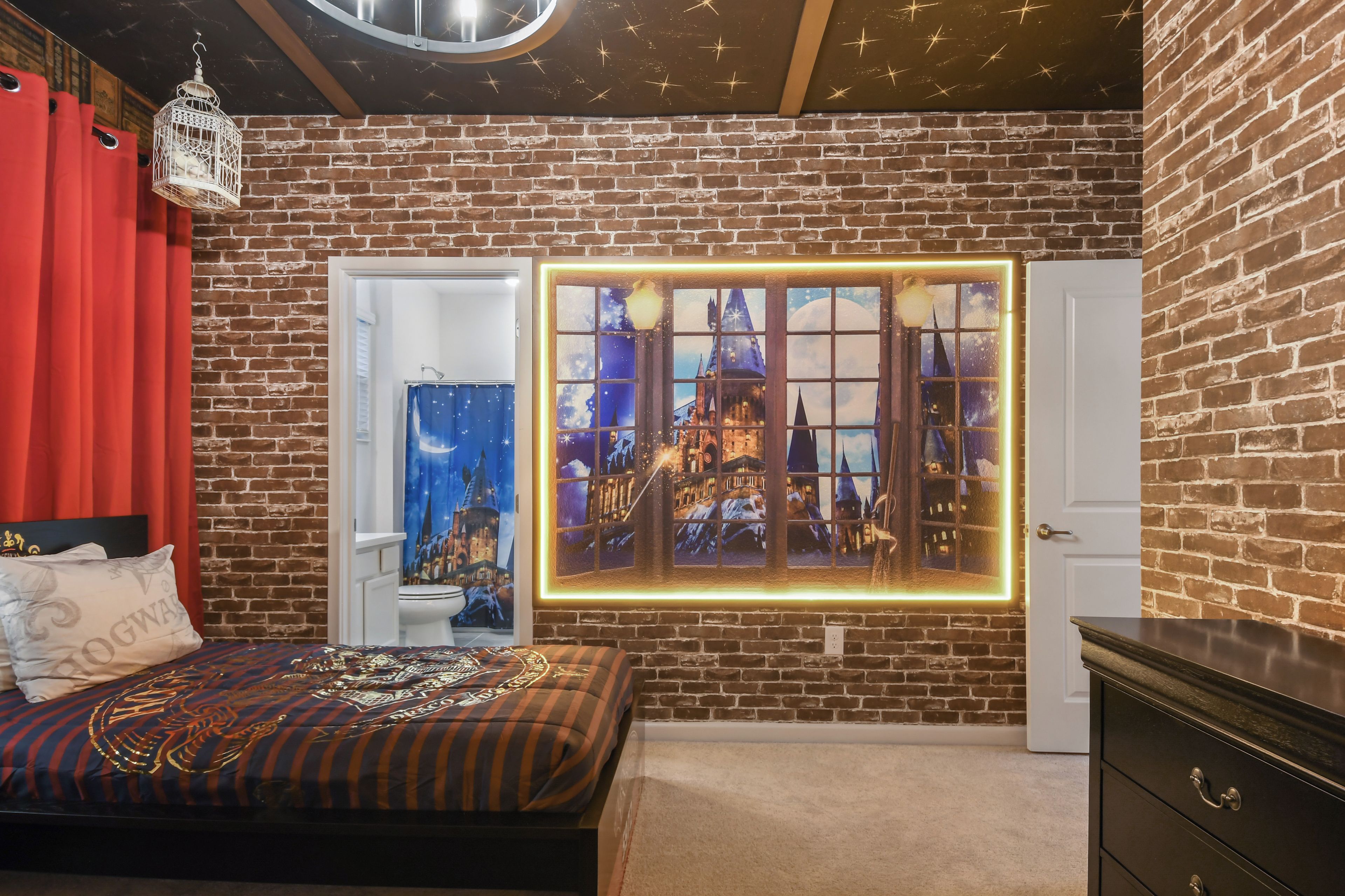 Ein Schlafzimmer mit Backsteinwand, einem Bett mit thematisch passender Bettdecke, einem Fenster mit Nachtsky-Motiv und einer Hängeleuchte.