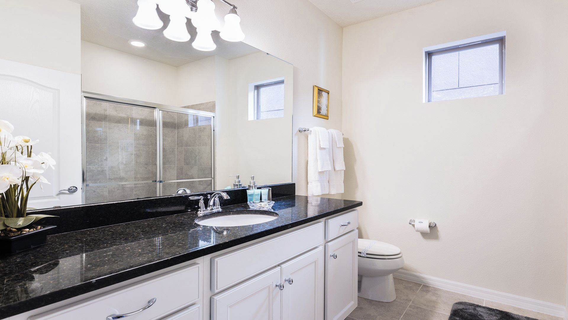 A bathroom with a large mirror, a black countertop, white cabinets, a toilet, and a small window.