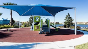 Un parque infantil con un toldo azul, suelo de goma roja y juegos de colores verde y amarillo.