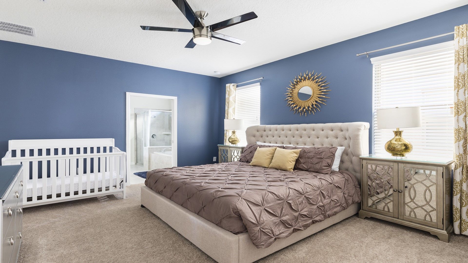 A bedroom with a bed, a crib, a ceiling fan, a vanity with a mirror, and a window with blinds.