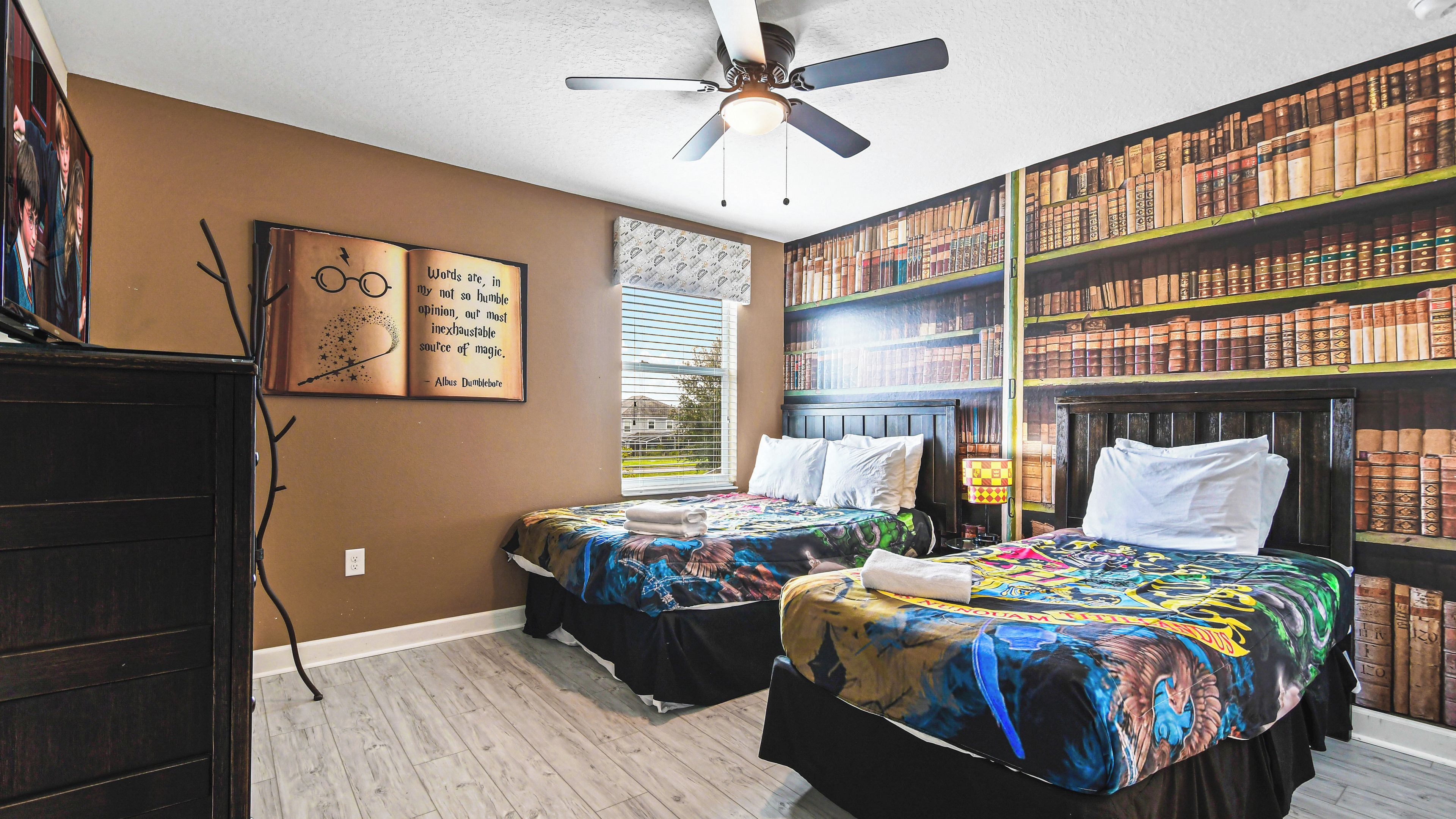 A bedroom with two beds, a ceiling fan, a bookshelf, and a window with blinds.