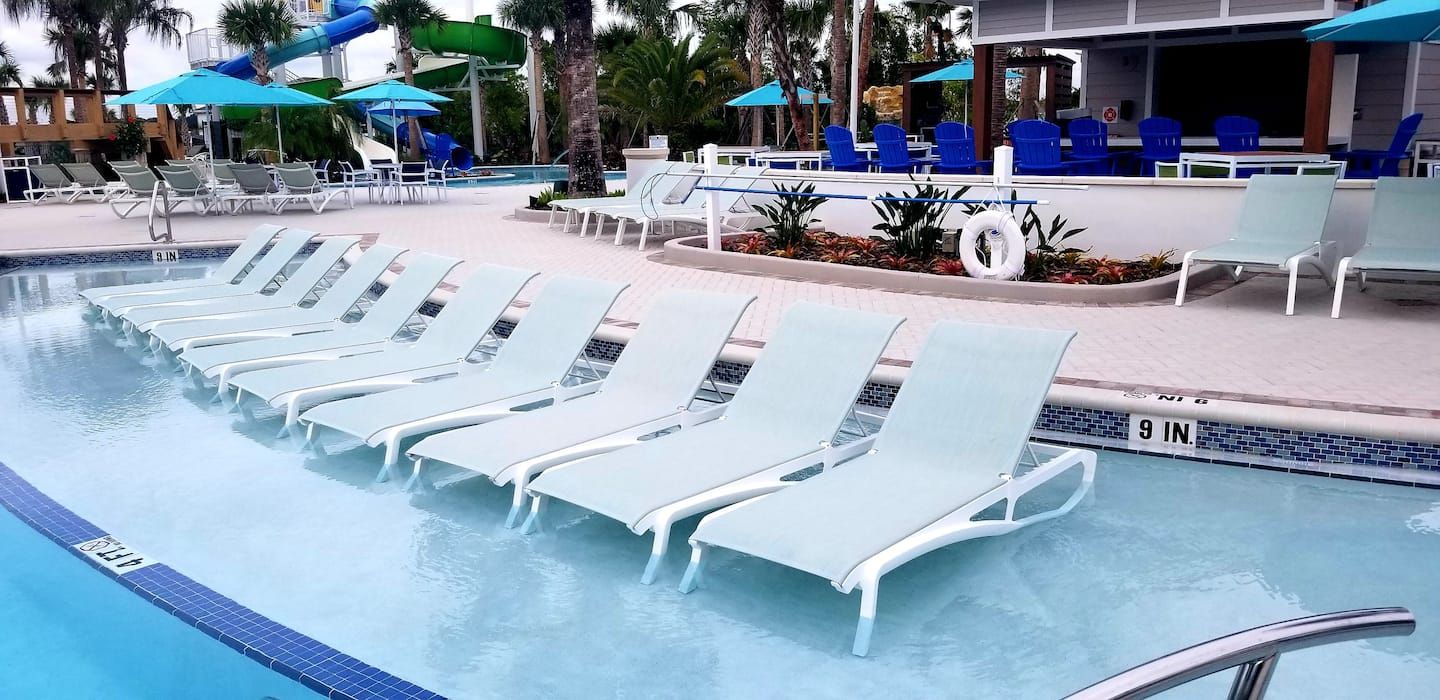 A pool area with white lounge chairs aligned along the edge, a water slide in the background, and blue umbrellas providing shade.