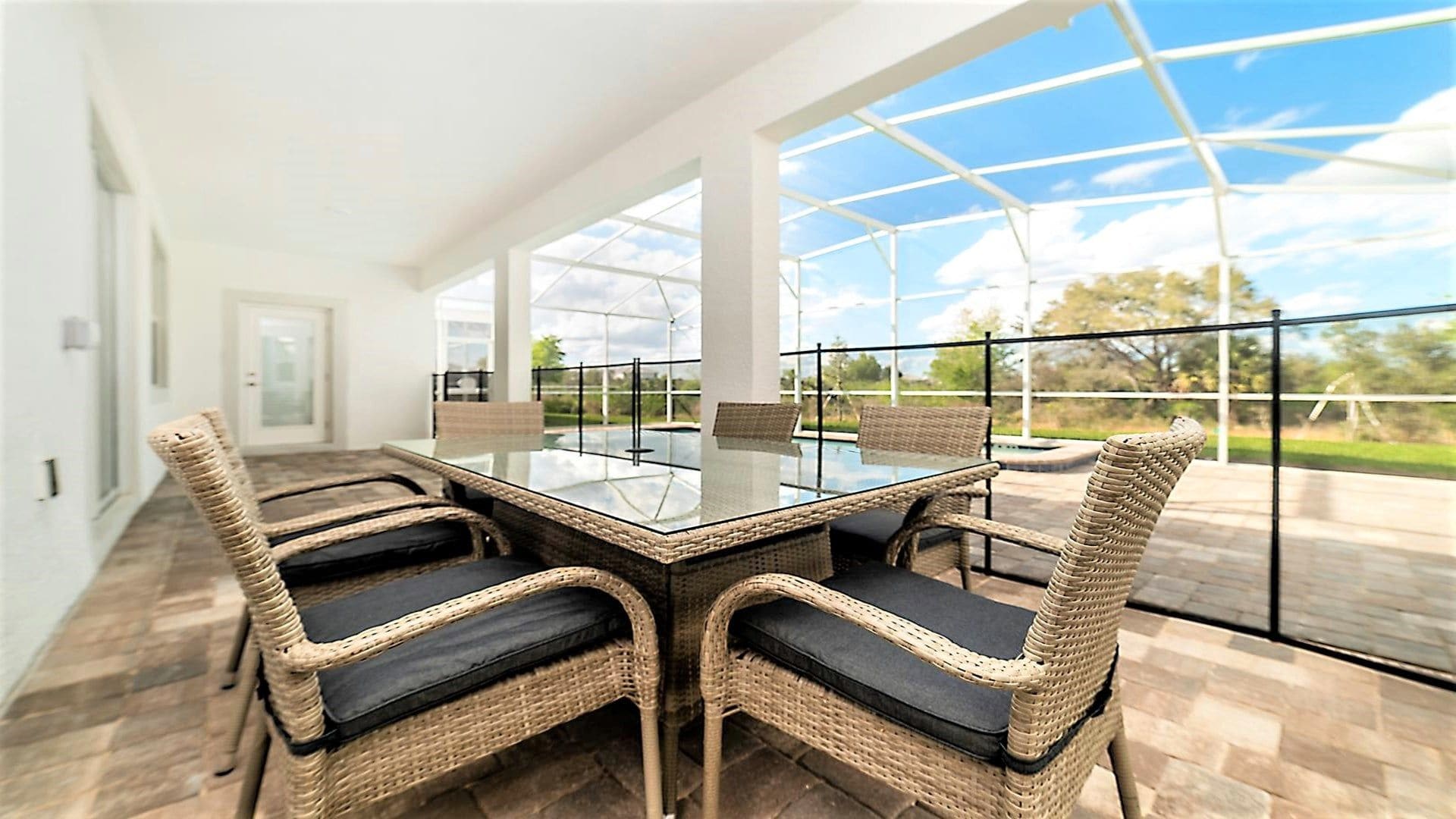 Un coin repas avec une table à plateau de verre entourée de chaises en osier dotées de coussins noirs, de grandes fenêtres et une vue sur l’extérieur.