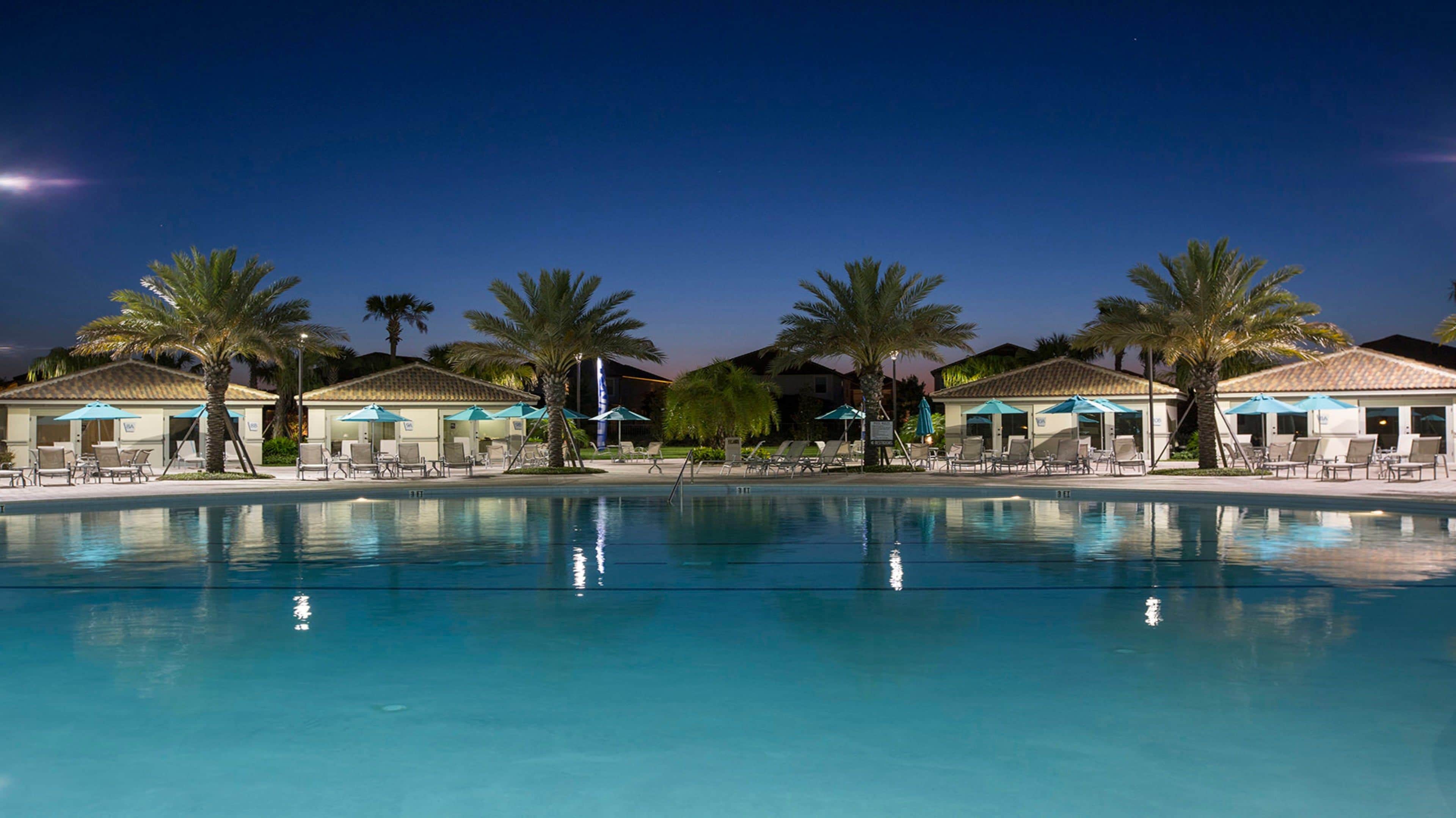 A swimming pool with lounge chairs and umbrellas, surrounded by palm trees and buildings with tiled roofs.