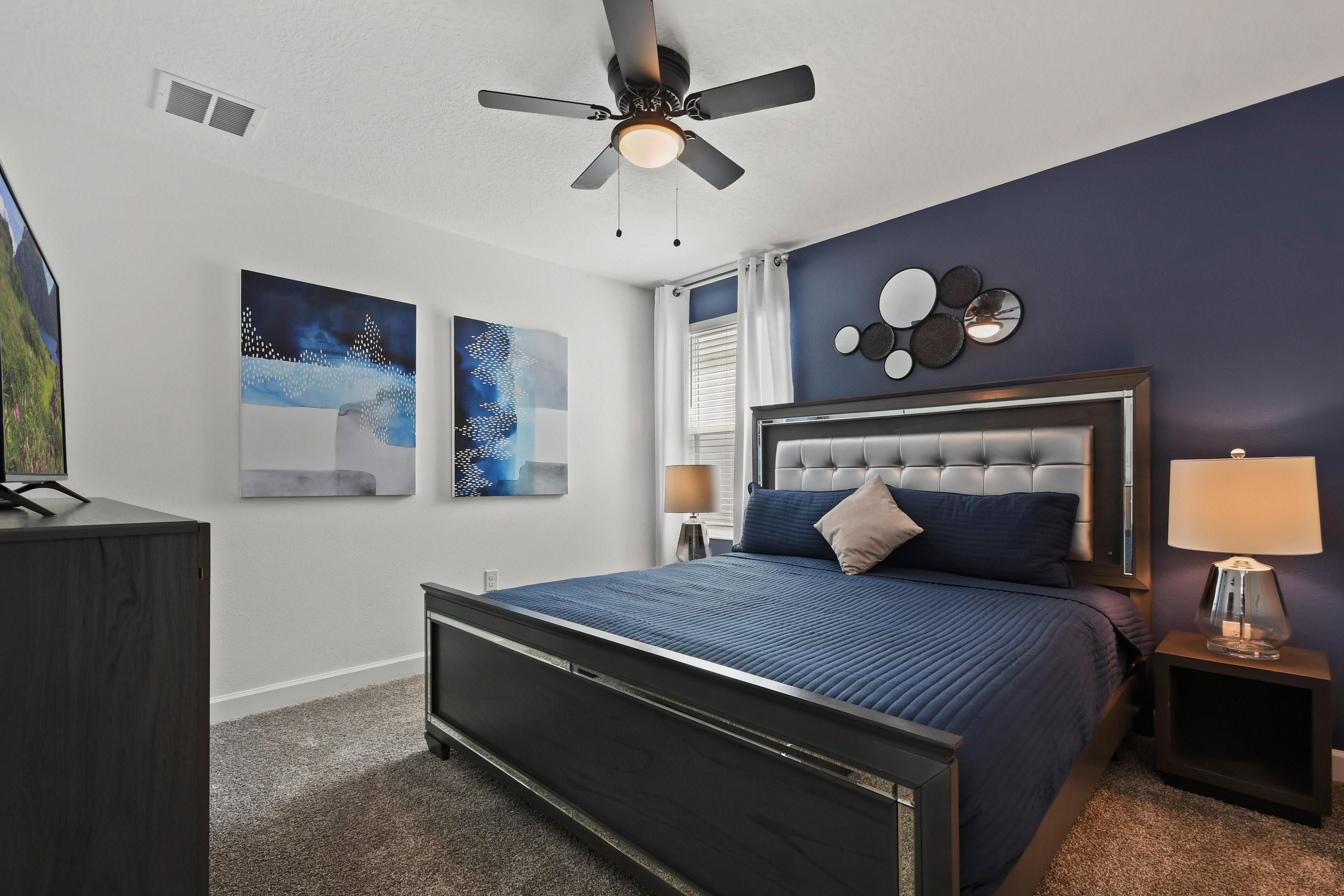 Une chambre à coucher avec un lit, un ventilateur de plafond, des œuvres d’art accrochées aux murs et une table de chevet avec une lampe.