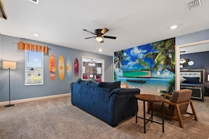 Ein Zimmer mit einem blauen Sofa, einem kleinen Tisch und einem Stuhl. Es gibt einen Deckenventilator, ein Fenster mit Jalousien und eine Wand mit Surfbrettern und einem Strandgemälde.