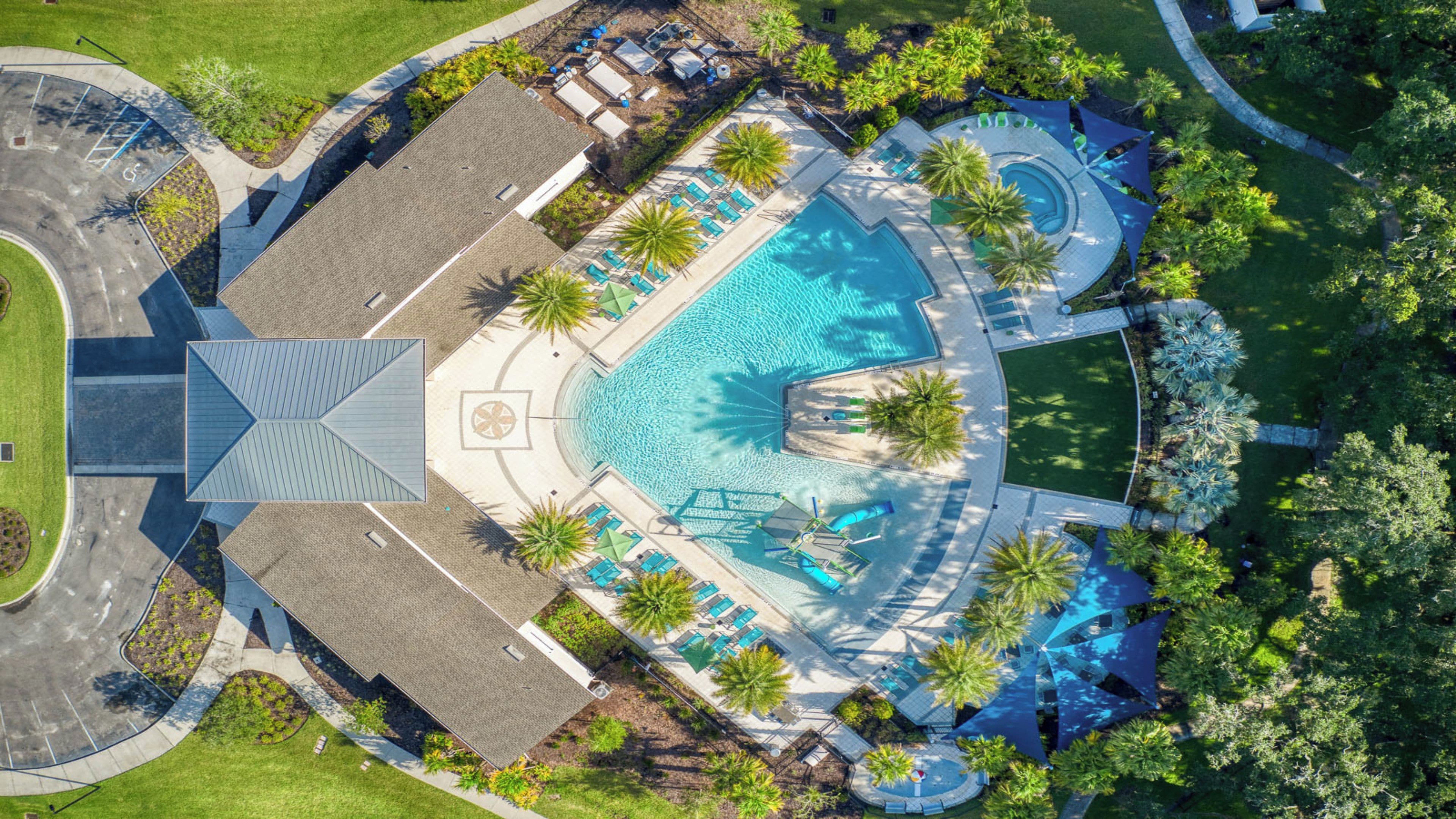An aerial view of a resort with a central swimming pool, multiple lounge areas, and palm trees.