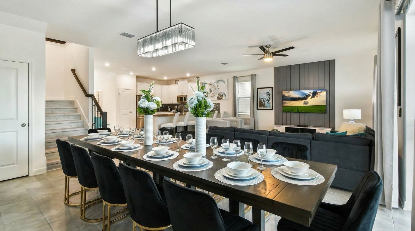 Un'area pranzo moderna con un tavolo lungo apparecchiato per sei persone, una scala, una cucina e un soggiorno con un divano e una TV.