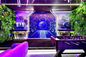 A room with a large digital screen displaying a futuristic tunnel, two foosball tables, and a bar area with a TV. - New! Avatar Game Room & Harry Potter Fantasy World (Davenport)
