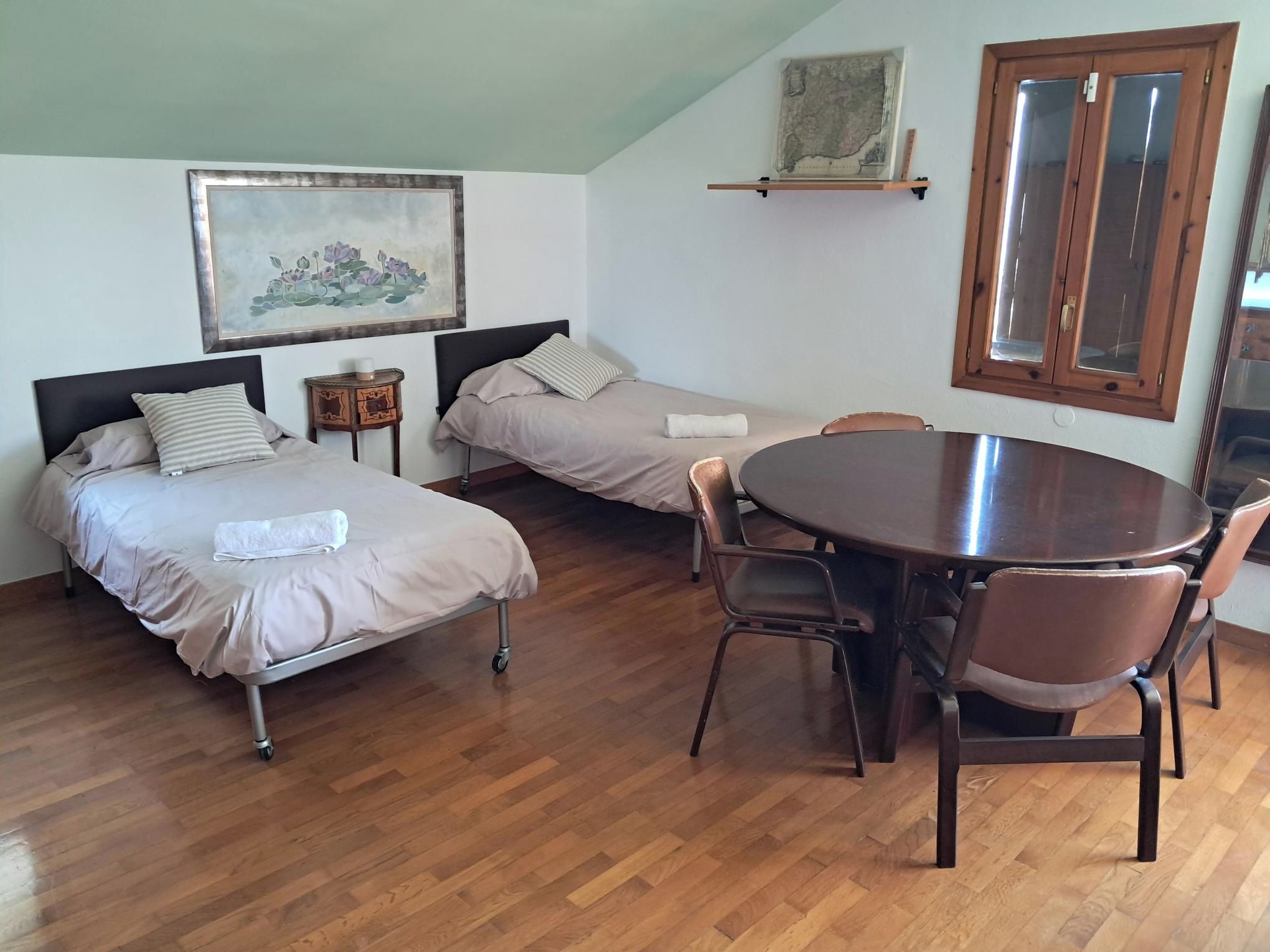 Une pièce avec deux lits, une table à manger avec des chaises, un plancher en bois, une carte accrochée au mur et une fenêtre avec un cadre en bois.