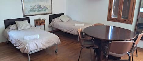 A room with two beds, a dining table with chairs, a wooden floor, a map on the wall, and a window with a wooden frame.