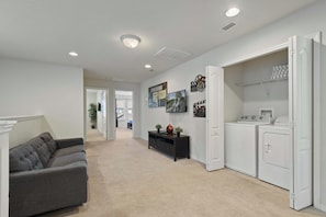 A modern living room with a grey sofa, a hallway leading to a bedroom, and a laundry area with a washing machine and dryer. - Cozy 5BR Townhome w/ Mickey Themed & Pool (Kissimmee)
