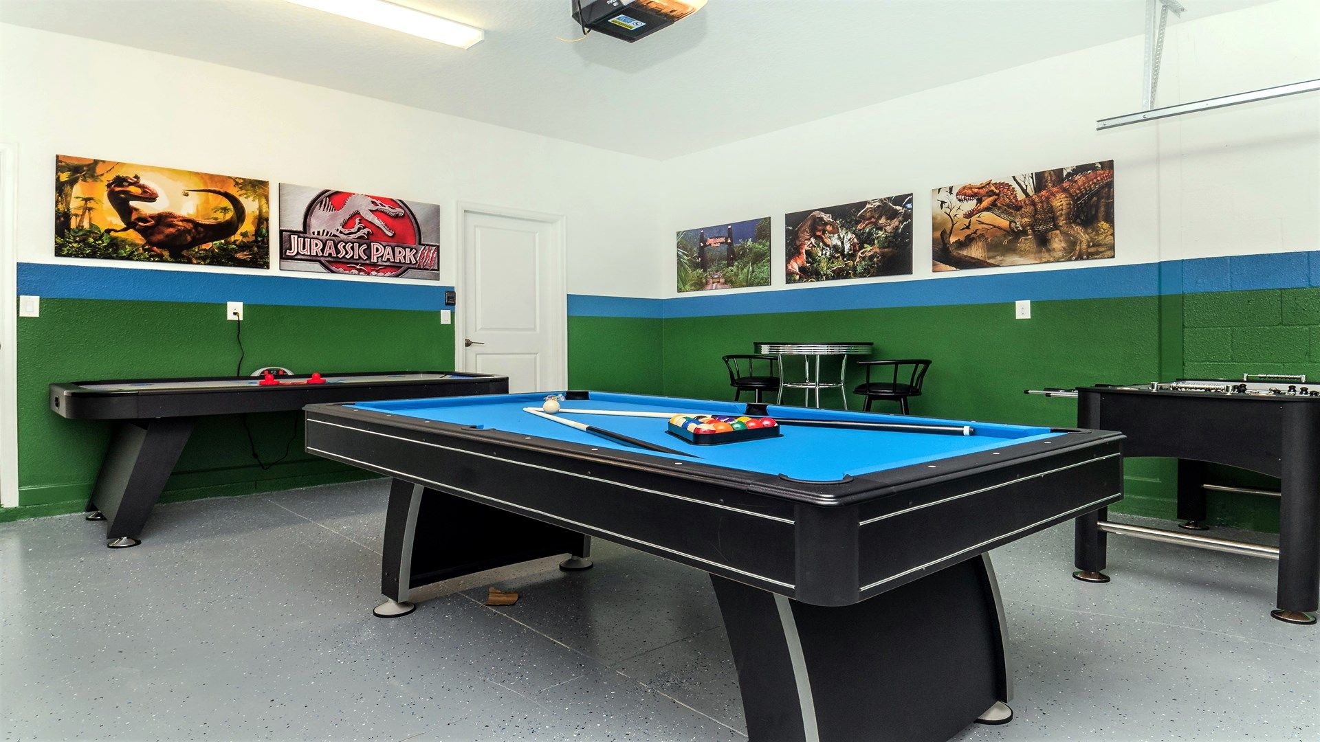 A pool table with a blue felt surface, surrounded by green walls with a white door. There are three posters on the wall, one of which is the 'Jurassic Park' logo.