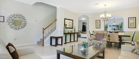 A spacious living room with a glass coffee table, a staircase, and a dining area with chairs and a chandelier.