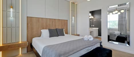 Une chambre à coucher moderne avec un grand lit, des tables de chevet, un banc, un miroir et une salle de bain visible au fond.