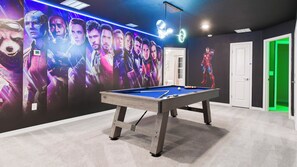 A pool table in a room with a wall mural of characters, a ceiling light, and a green-lit door. - Massive 10br Villa! Themed Bunks & Waterpark (Davenport)