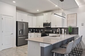 Une cuisine moderne avec des meubles blancs, des appareils en acier inoxydable, un grand îlot central doté de tabourets et un dosseret noir en carreaux.