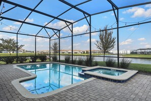 A swimming pool with a hot tub, surrounded by a covered area with a view of a lake and buildings in the background. - Lakeview Home w/ Mario Game Room & Themed Bedrooms (Kissimmee)