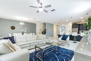 A modern living room with a white sofa, a glass coffee table, and a blue rug. There is a kitchen area with dark cabinets and a white countertop. - Championsgate Private Pool Spacious Luxury Home (Davenport)
