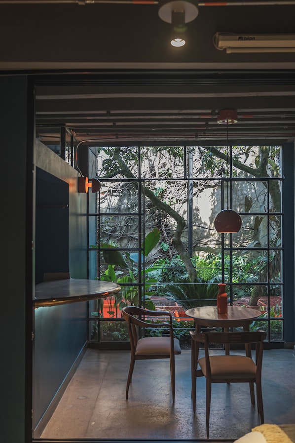 Una habitación con una mesa y sillas, una ventana con vista a follaje, y una lámpara en el techo.