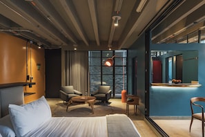Una habitación de hotel moderna con una cama, dos sillones, una mesita, un escritorio con un frutero y un ventanal con vistas.