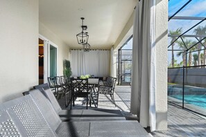 A covered outdoor patio with a table and chairs, a hanging light fixture, and a view of a swimming pool. - Family Fun Villa: Themed Rooms, Private Pool & Spa (Clermont)