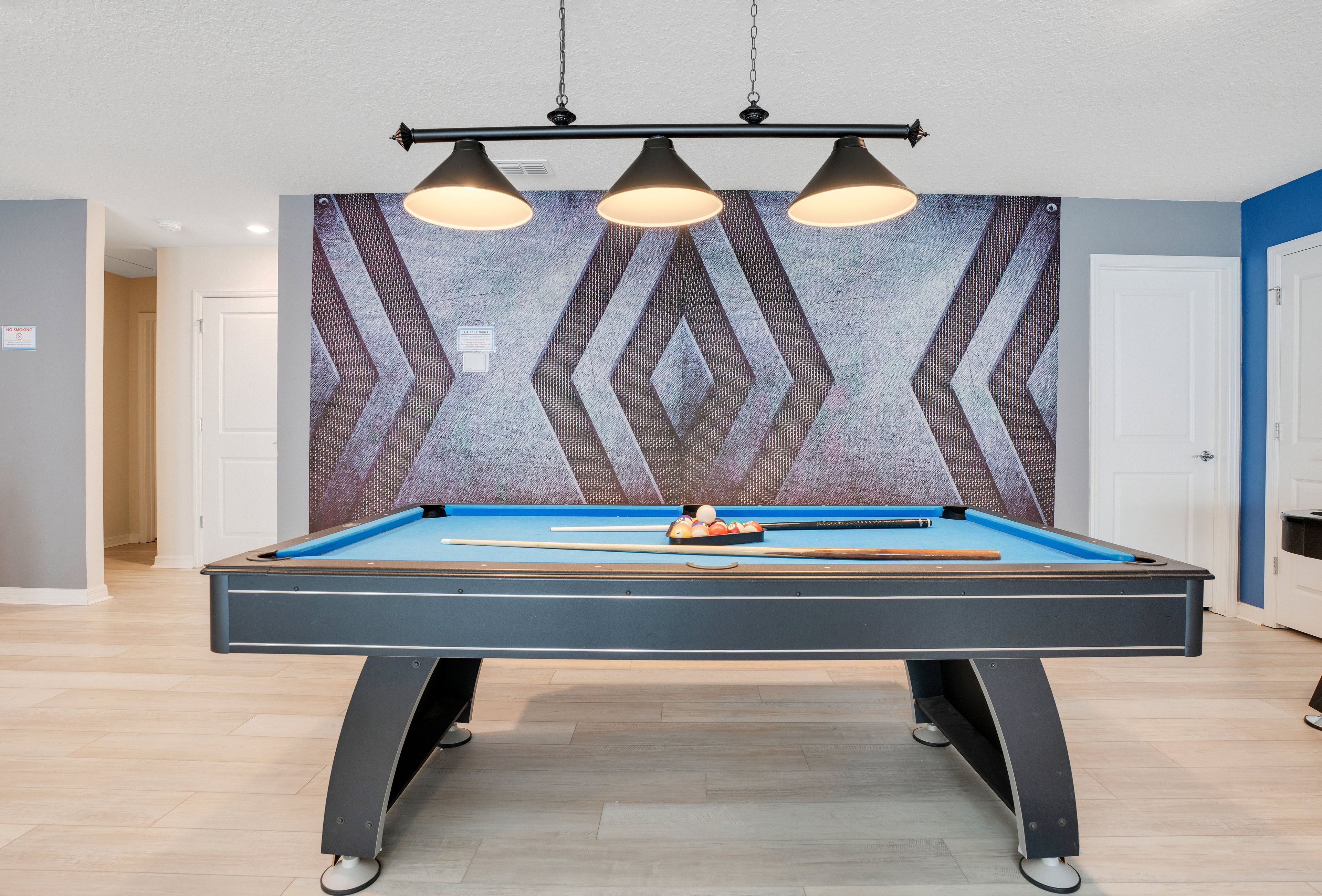 A pool table with a geometric wall mural, three pendant lights, and a pool cue rack.