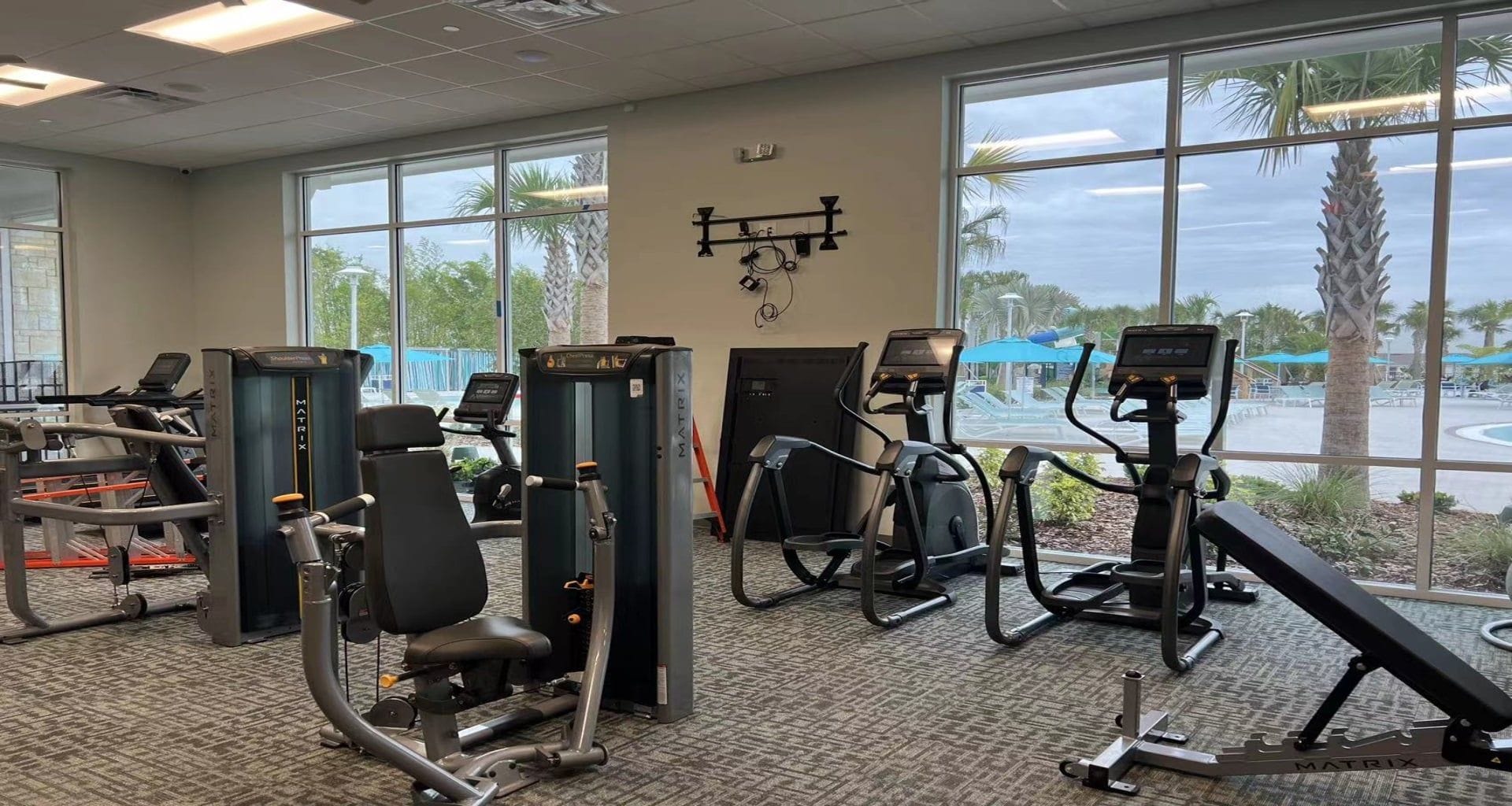 Ein Fitnessstudio mit unterschiedlichen Trainingsgeräten, großen Fenstern und Blick auf Palmen.