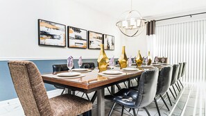 Ein Esszimmer mit einem langen Tisch, gedeckt für sechs Personen, ausgestattet mit einem modernen Kronleuchter, abstrakter Wandkunst und Blick ins Freie durch große Fenster.