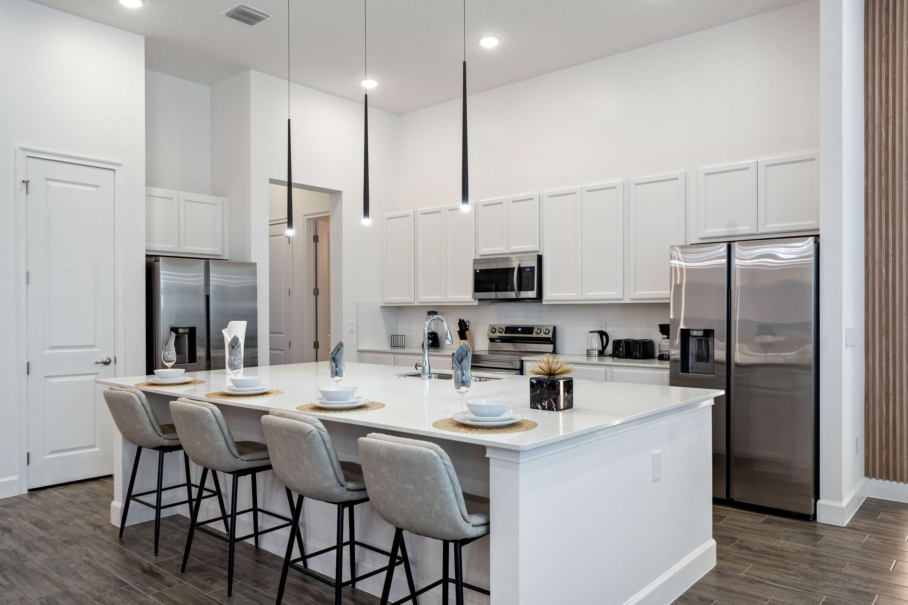 Cocina moderna con muebles blancos, electrodomésticos de acero inoxidable, una isla central con taburetes altos, iluminación colgante y un refrigerador grande.