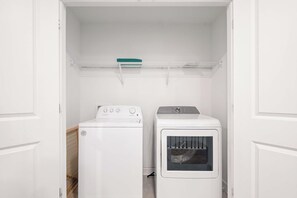 A laundry room with a front-loading washer and dryer, a shelf above the dryer, and a door leading to another room. - Games/themed Rooms/greenview/ 9BR @ Storeylake! (Kissimmee)