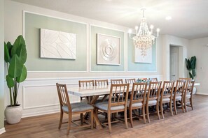 A dining area with a long table and wooden chairs, flanked by large potted plants and framed artwork on the walls. - Resort Access/7br Family-villa/close2parks! (Clermont)