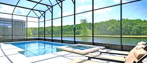 Une piscine entourée de chaises longues, le tout protégé par une structure en verre offrant une vue sur un lac et une forêt.