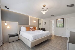 Une chambre moderne avec un grand lit, des tables de chevet, une commode, un luminaire au plafond et une porte donnant sur une autre pièce.