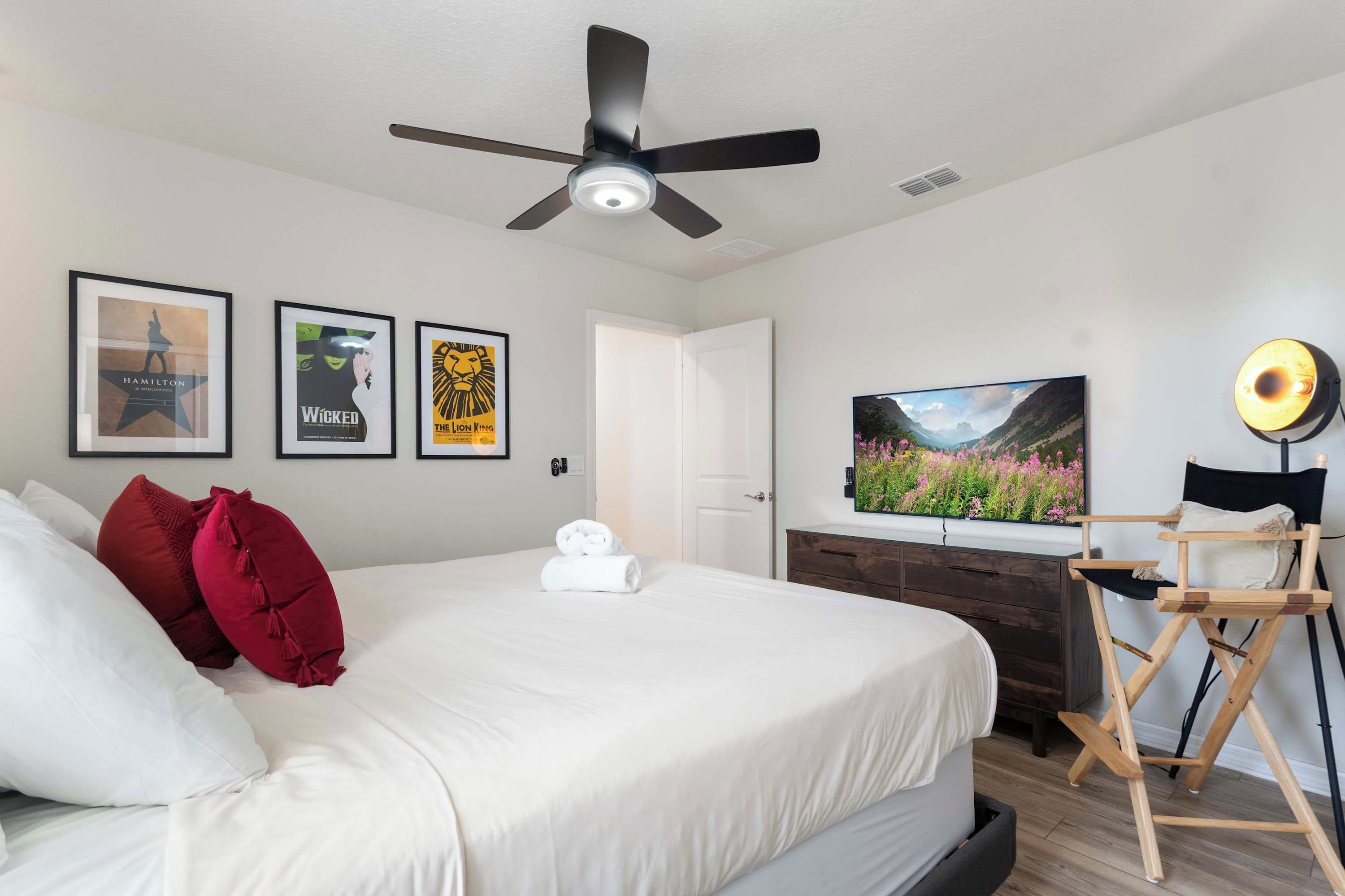 A hotel room with a bed, ceiling fan, framed posters, a television, and a director's chair.
