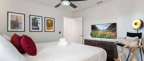 A hotel room with a bed, ceiling fan, framed posters, a television, and a director's chair.