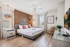 Ein modernes Schlafzimmer mit Bett, Nachttischen, Deckenventilator, einem Fenster mit Vorhängen, einem an der Wand montierten Fernseher und einem Gemälde.