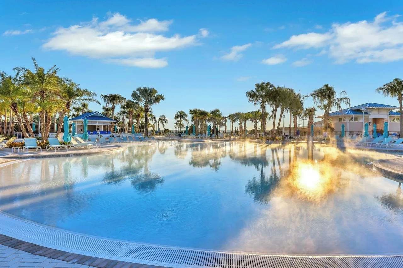 Piscina com cadeiras de descanso e palmeiras, cercada por edifícios com telhados azuis.