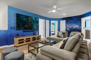 A living room with a blue wall, a large flat-screen TV, a wooden cabinet, a beige sofa, and a glass coffee table. - Themed Villa, Loaded Games w Pool Near Parks! (Davenport)