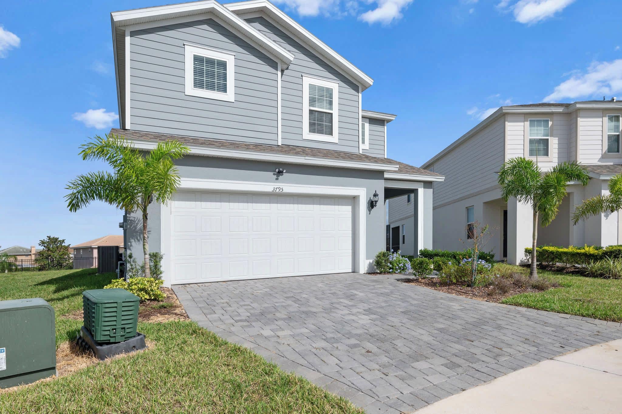 A two-story residential house with a white garage door, a paved driveway, and a small front yard with a palm tree and shrubs.