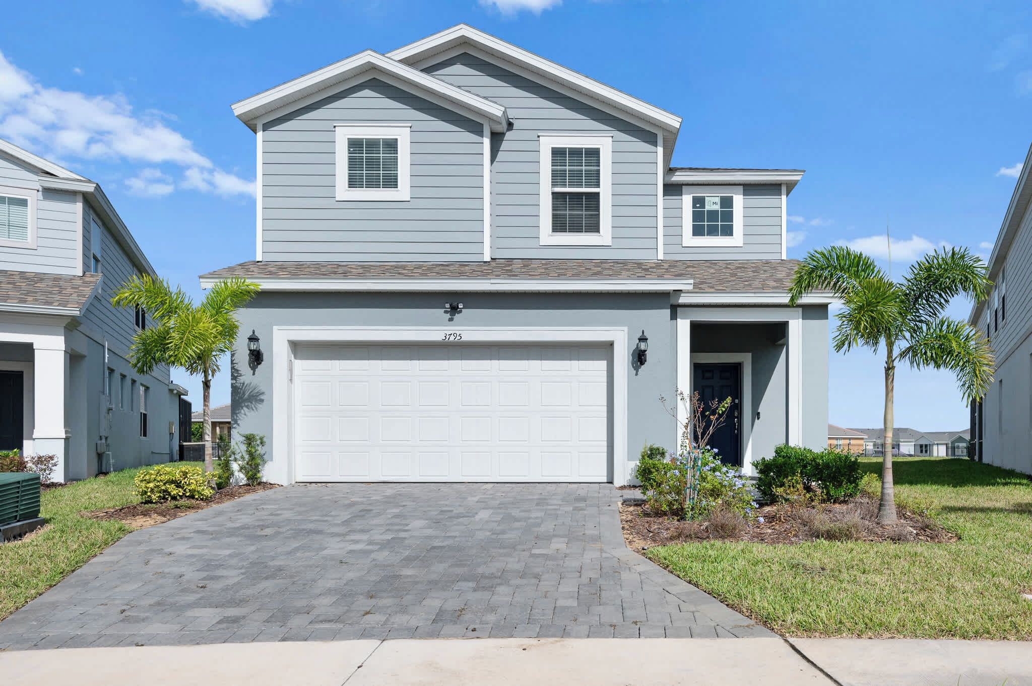 A two-story residential house with a garage, a driveway, and a well-maintained lawn with palm trees.