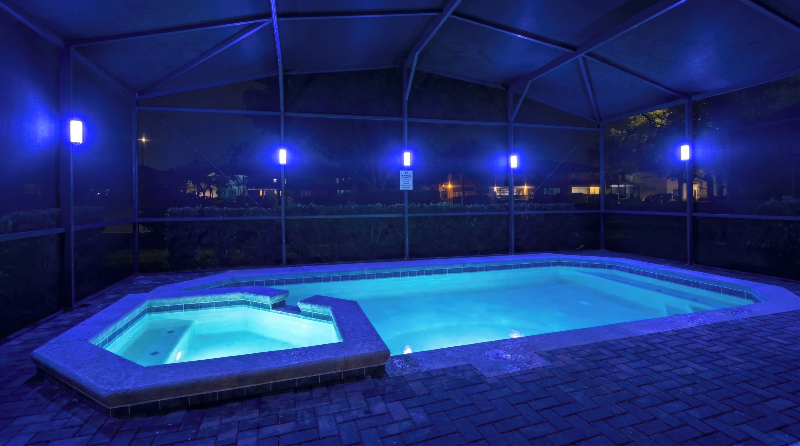 A heated outdoor swimming pool with a glass enclosure, surrounded by a brick patio.
