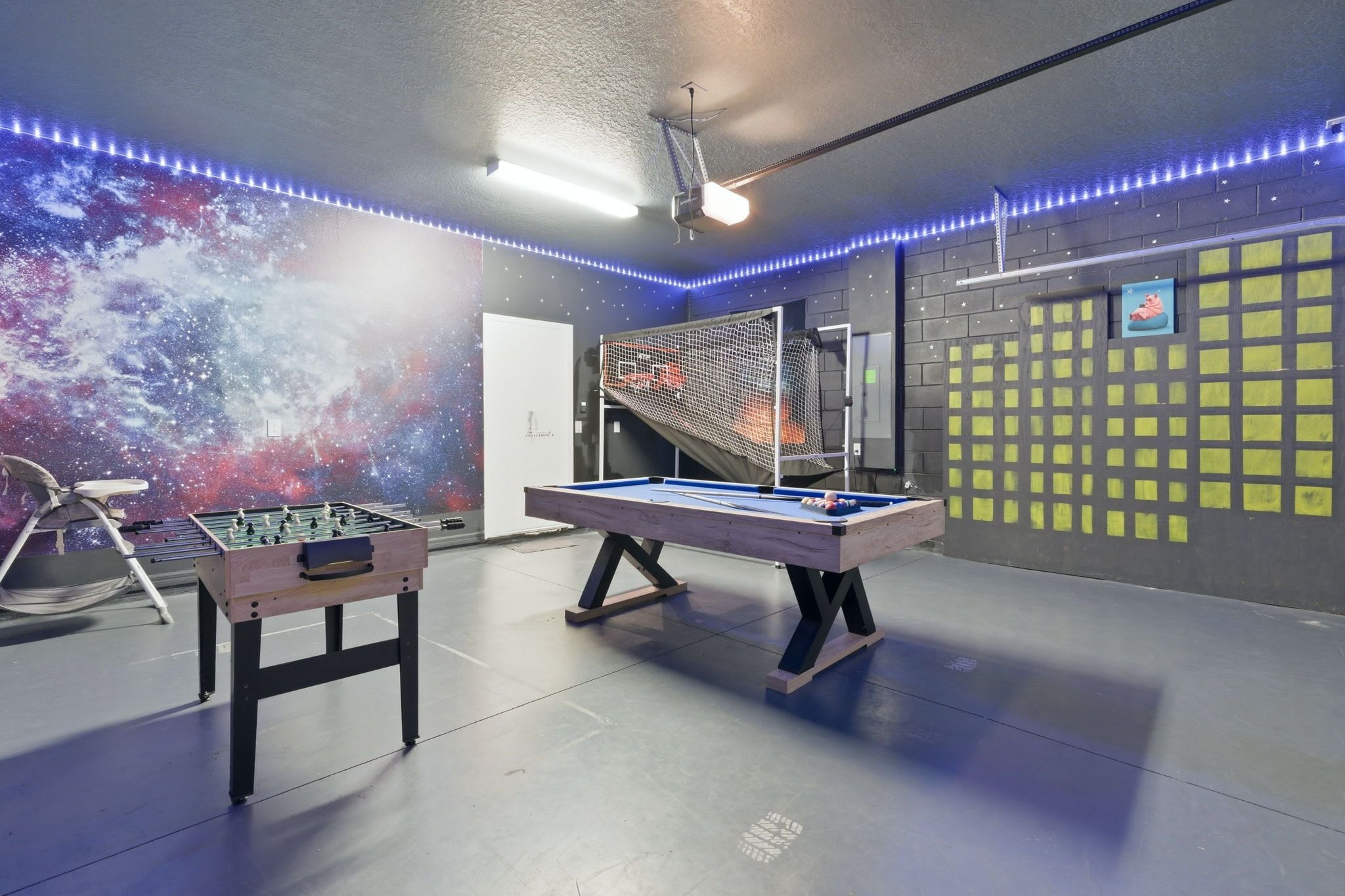 A foosball table, a pool table, and a foosball table in a room with a galaxy-themed wall and LED lights.