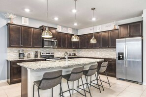 A modern kitchen with dark wood cabinets, stainless steel appliances, a granite island with bar stools, pendant lighting, and a tiled backsplash. - Themed Family Retreat With Epic Game Room (Davenport)