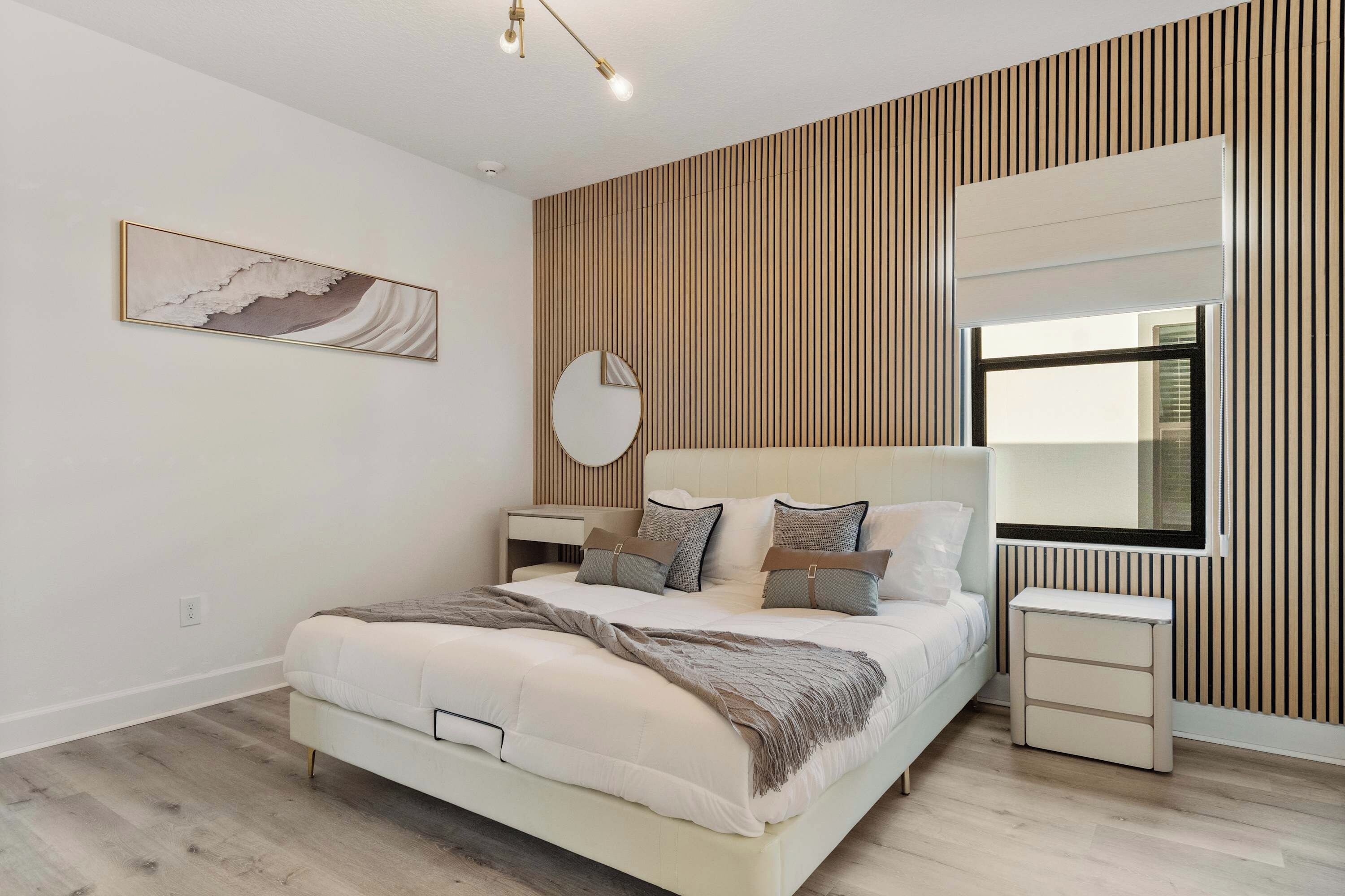Un dormitorio moderno con una cama grande, mesitas de noche, un espejo redondo y una ventana con persianas.