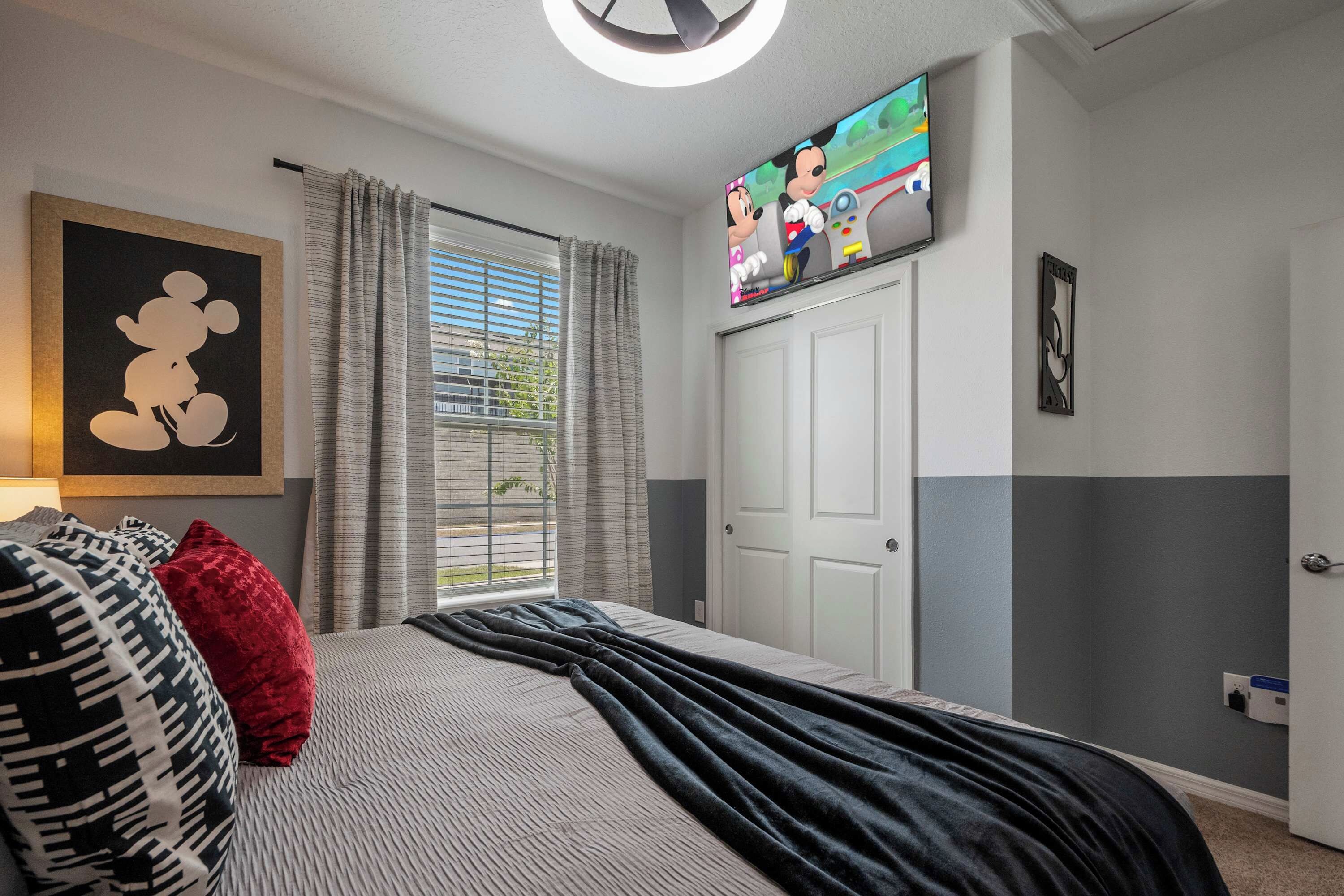 Un dormitorio con una cama, un cuadro de Mickey Mouse, un televisor y una ventana con cortinas.