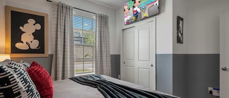 Un dormitorio con una cama, un cuadro de Mickey Mouse, un televisor y una ventana con cortinas.