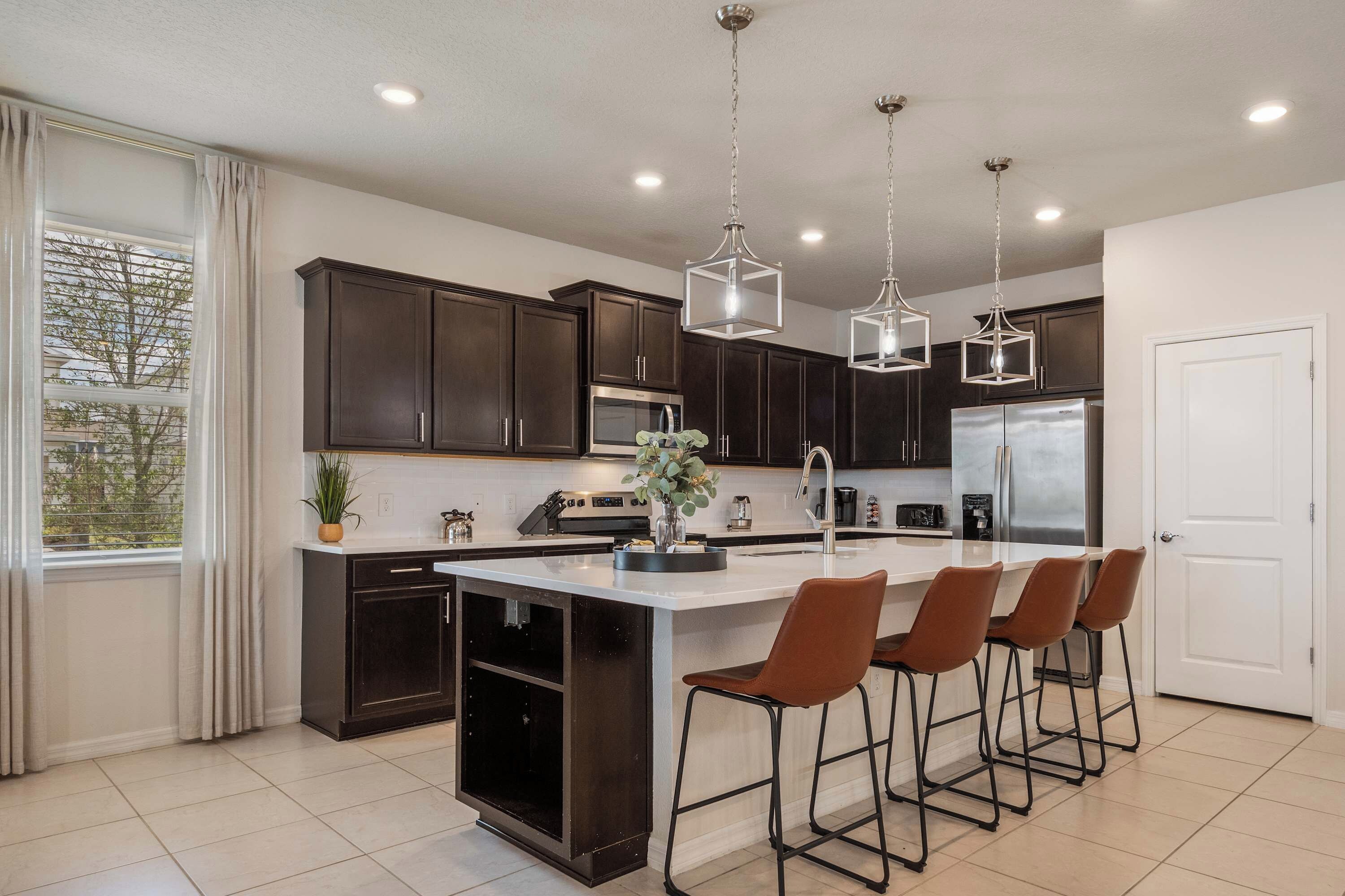 A modern kitchen with dark cabinets, a central island with bar stools, pendant lighting, and a view through a window.